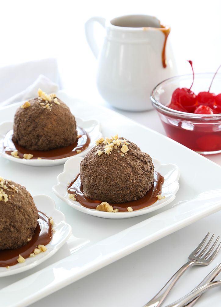 three desserts on plates with chocolate sauce and cherries in the bowl behind them