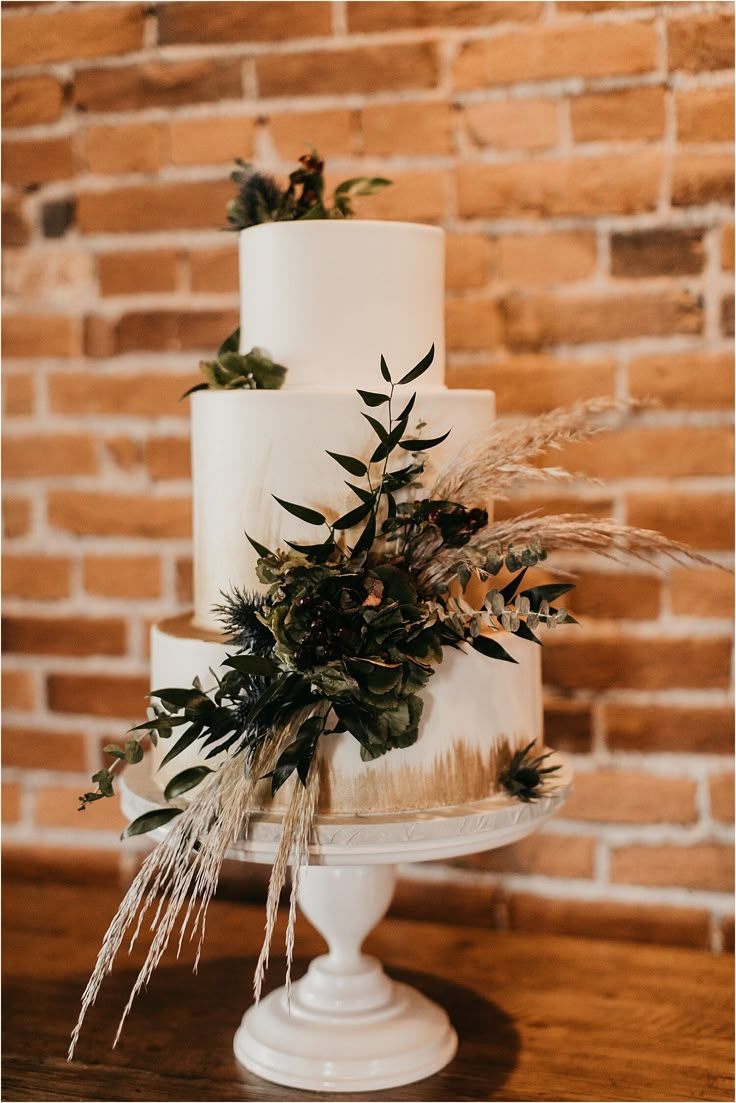 a white wedding cake with greenery on the top and bottom is sitting on a table in front of a brick wall