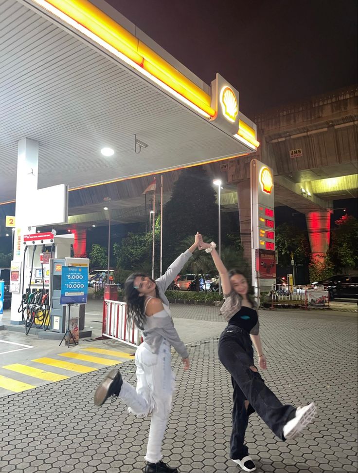 two people are dancing in front of a gas station with their arms up and one person is holding the other's hand