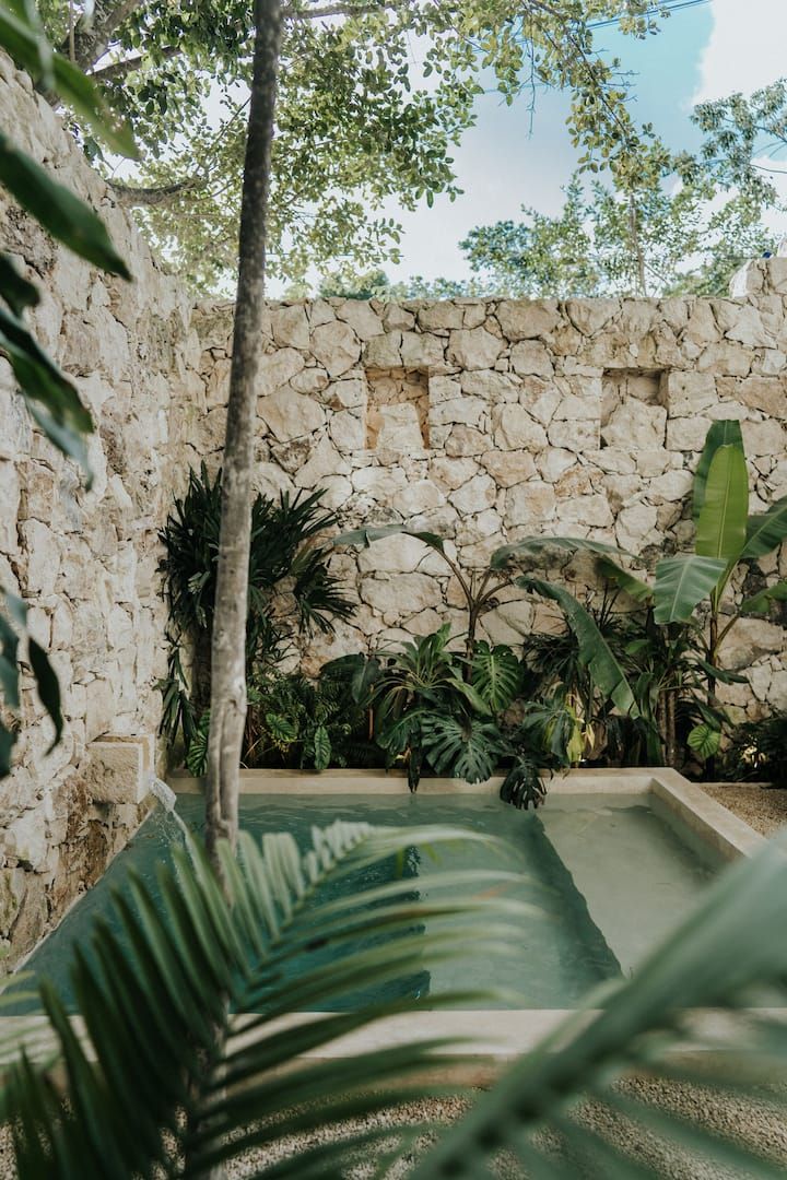 an outdoor pool surrounded by trees and plants in front of a stone wall with green foliage