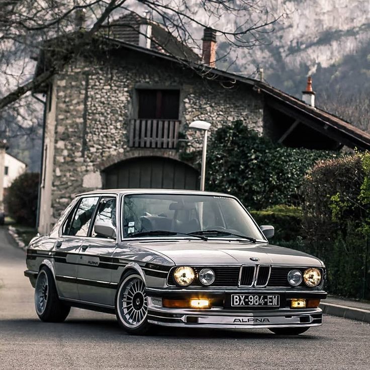 an old bmw is parked in front of a stone house with mountains in the background