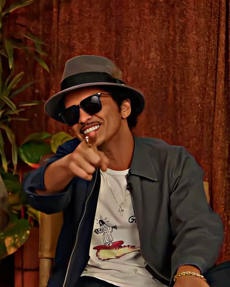 a man in a hat and sunglasses sitting down with his finger pointing at the camera