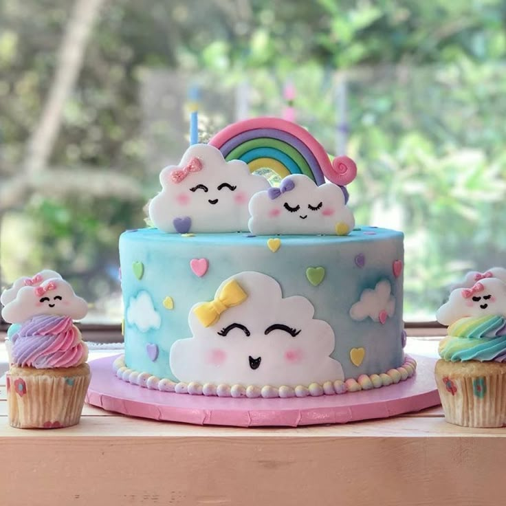 there is a cake that has been decorated with clouds and rainbows on it, as well as cupcakes