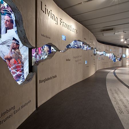 a large wall with pictures on it and people walking by in the hallway behind it