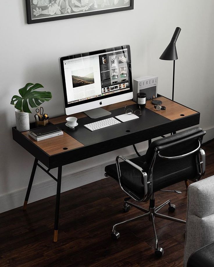 a desk with a computer on it in front of a chair and a painting hanging over the wall