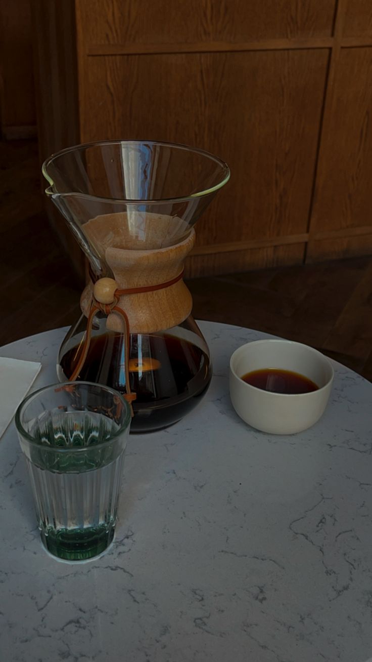 a table topped with a glass cup filled with liquid next to a coffee pot and saucer