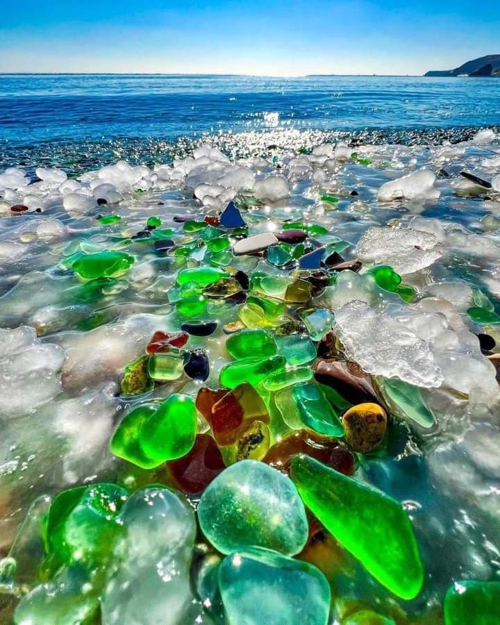 many different colored glass pieces are on the sand by the water's edge, and there is no image here to provide a caption