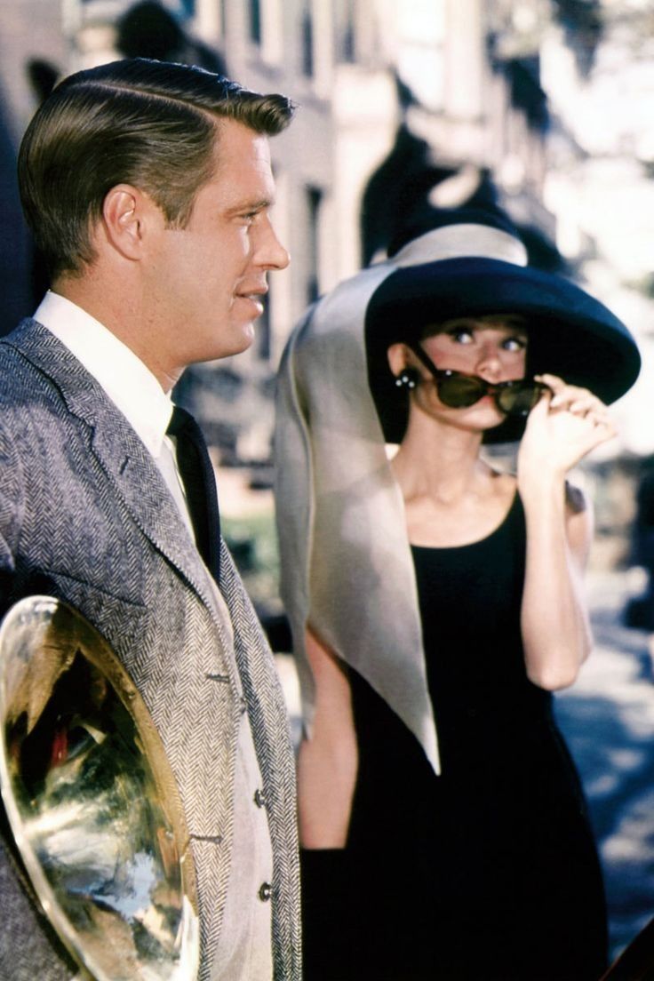 a man standing next to a woman in a black dress and hat on the street