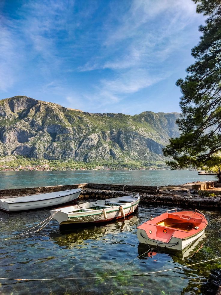 three small boats tied to the shore in front of a mountain range and clear water