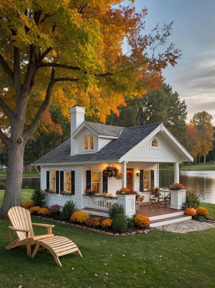 a small white house sitting next to a tree with lots of leaves on the ground