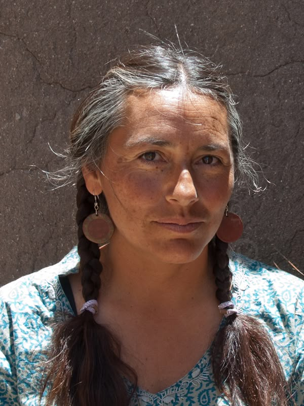 a woman with braids and earrings standing in front of a stone wall looking at the camera