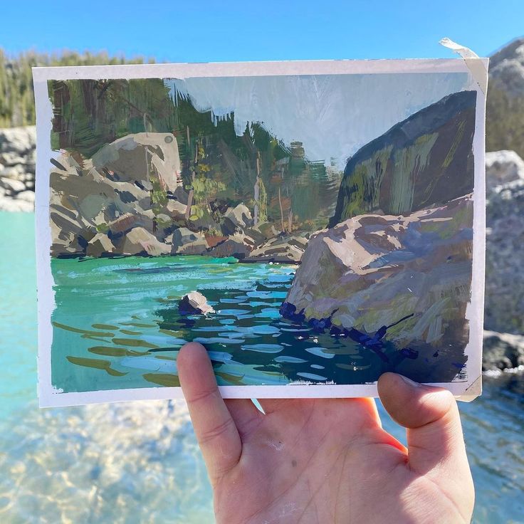 a hand holding up an image of a lake with rocks and trees in the background