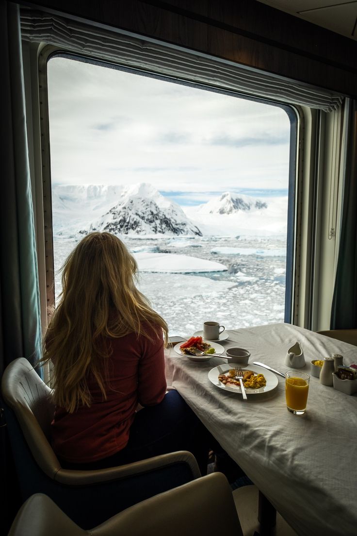 a woman sitting at a table with food and drinks in front of her looking out the window