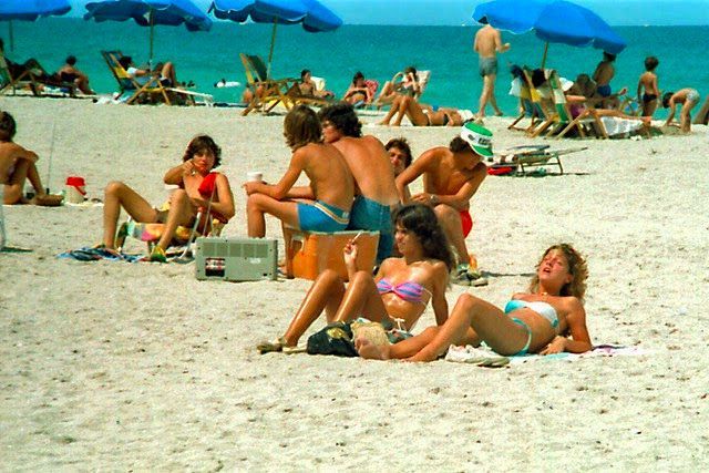 many people are on the beach with blue umbrellas in the background and one person is laying down