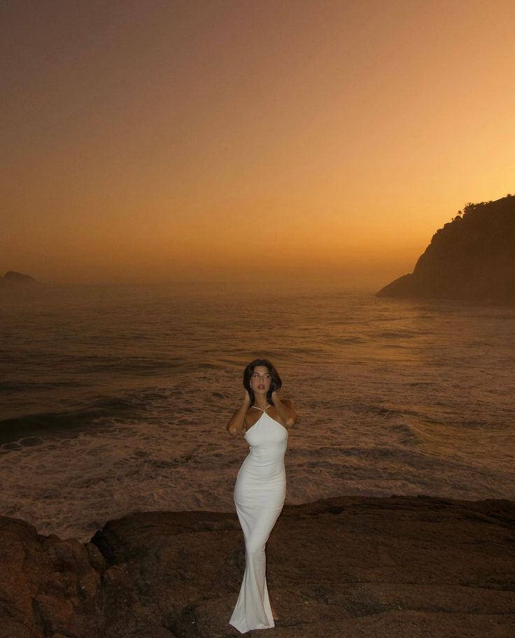 a woman standing on top of a rocky beach next to the ocean at sun set