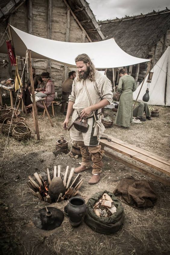 a man with long hair and beard standing next to an open fire in front of a tent