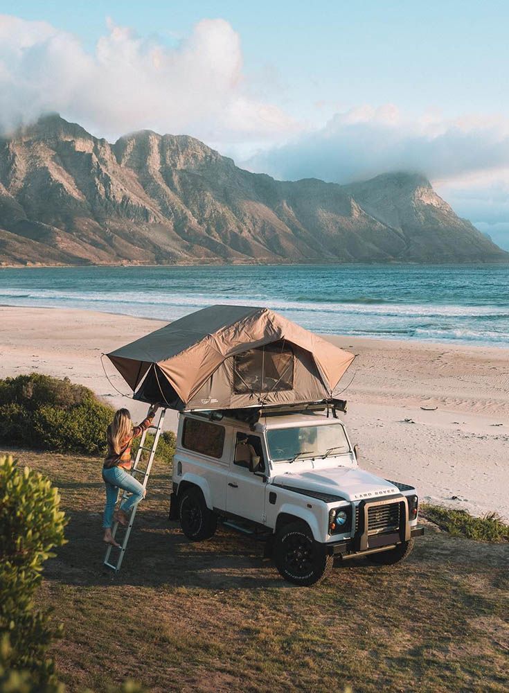 a woman standing next to a tent on top of a white truck near the ocean