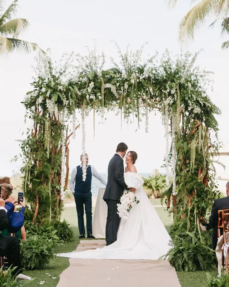 a bride and groom standing under an arch with greenery at their wedding ceremony on the lawn