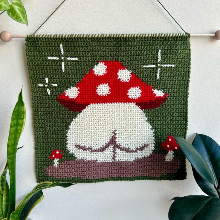 a green wall hanging with a red and white mushroom on it's side next to a potted plant