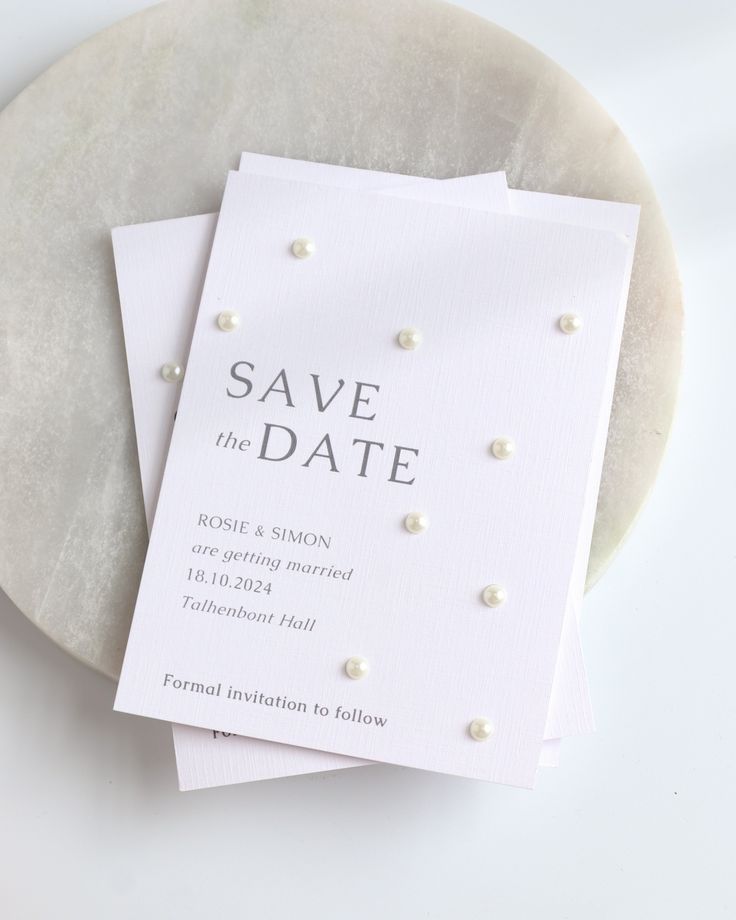 two wedding save the dates cards sitting on top of a white plate with silver pearls