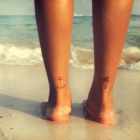 a woman's feet with a small anchor tattoo on her left foot, standing in the sand at the beach