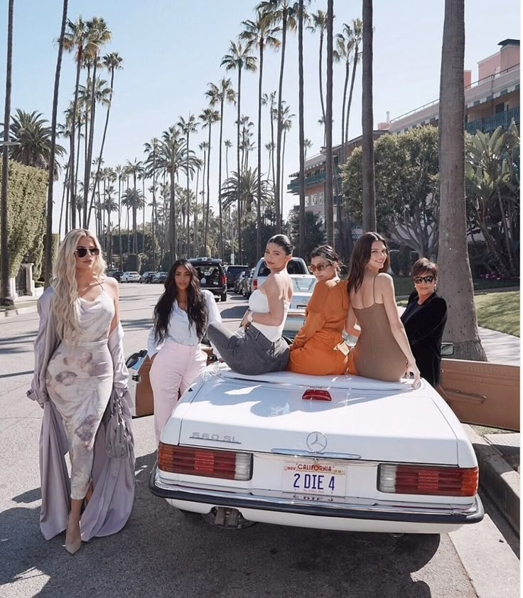 four women are sitting on the hood of a white car in front of palm trees