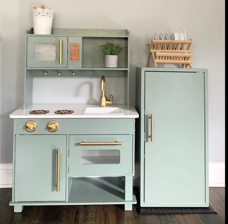 a green toy kitchen with sink and cupboards on the wall next to a wooden floor