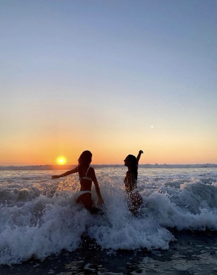 two people in the ocean at sunset with their arms out and one person on surfboard