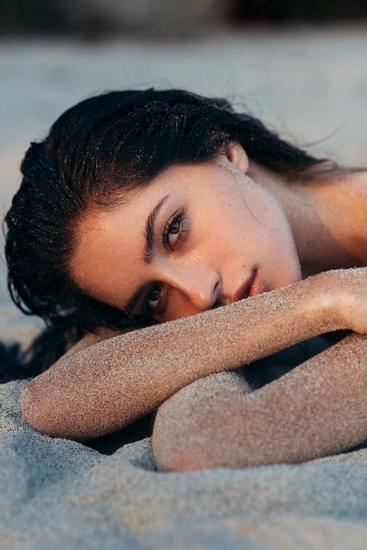 a beautiful woman laying on top of a sandy beach next to the ocean with her eyes closed