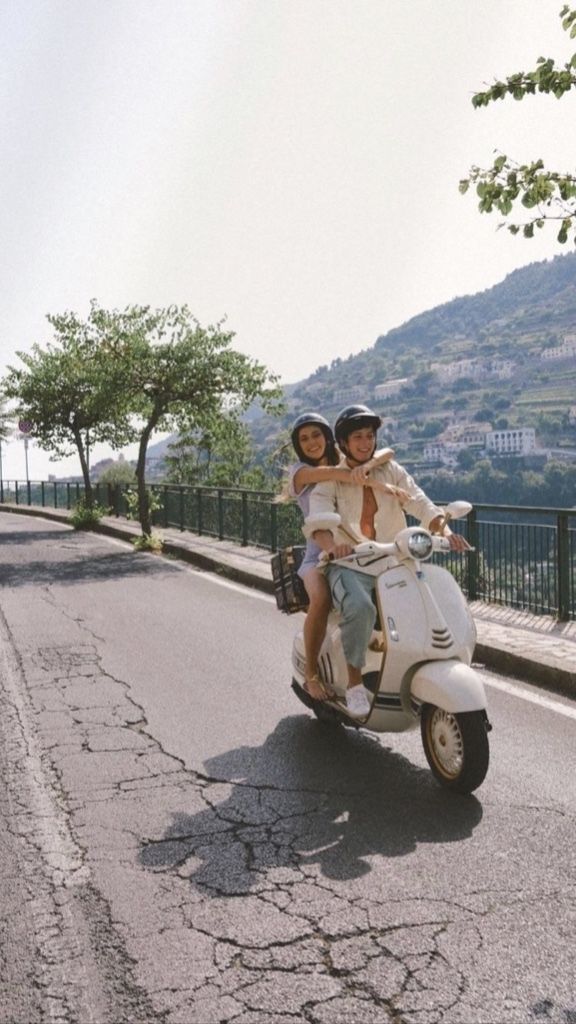 two people riding on the back of a scooter down a road next to trees