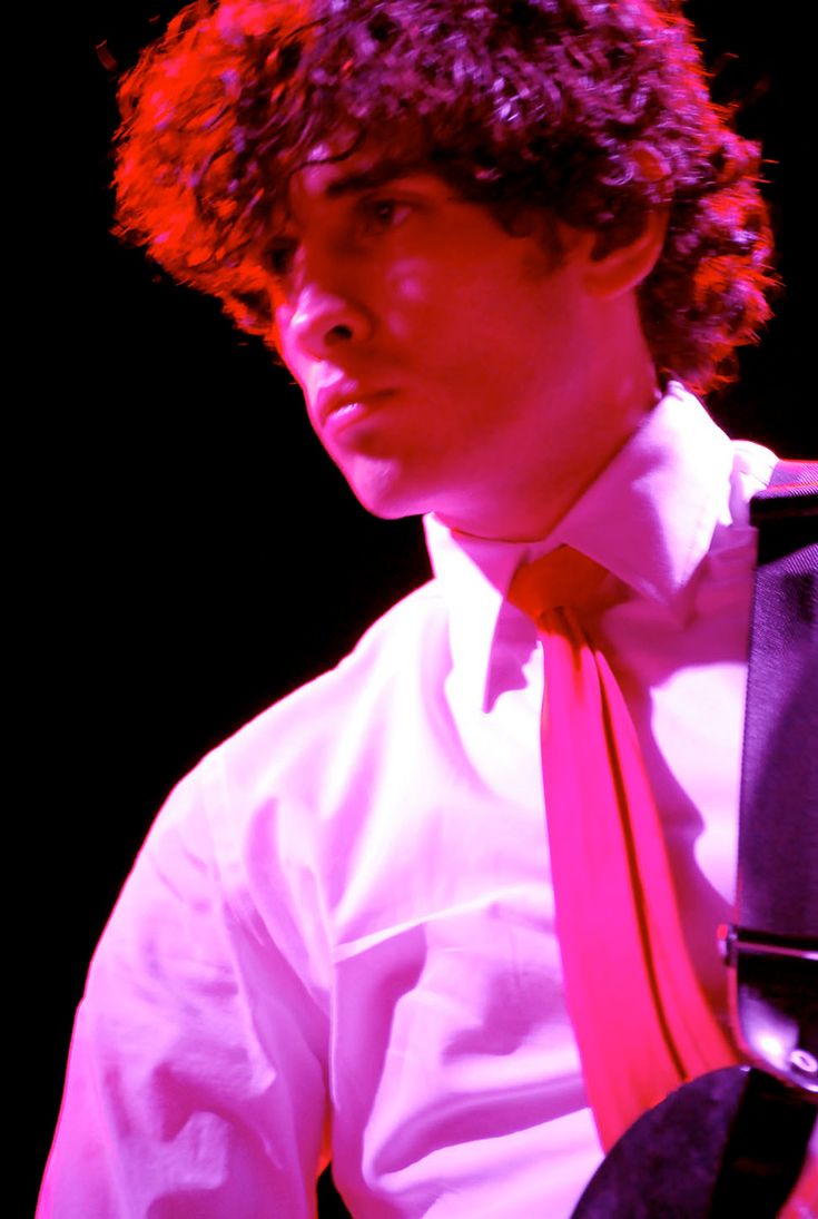 a young man with curly hair wearing a white shirt and pink tie, standing in front of a black background