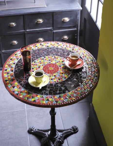 two cups of coffee sit on a mosaic table in front of an ornate dresser and window