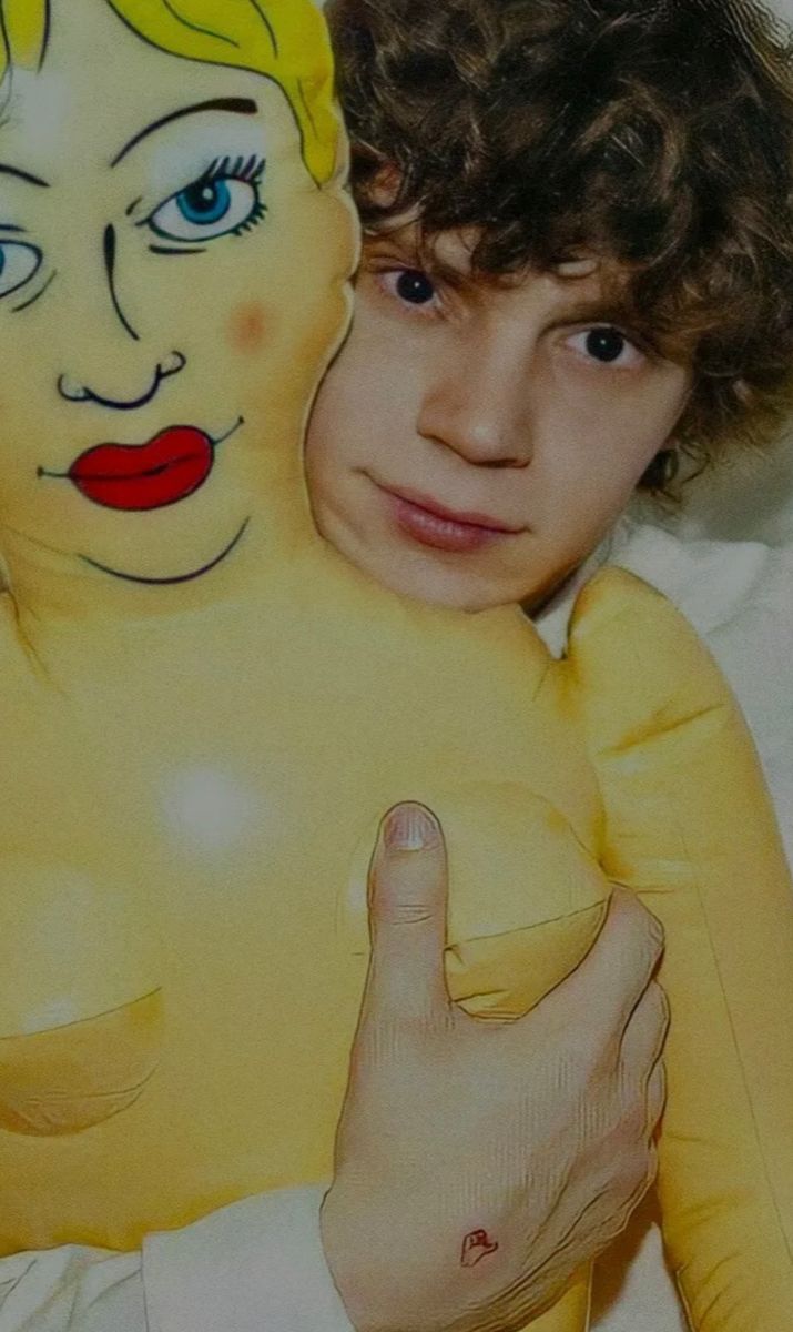 a young man with curly hair and blue eyes is holding a yellow balloon shaped like a woman's face