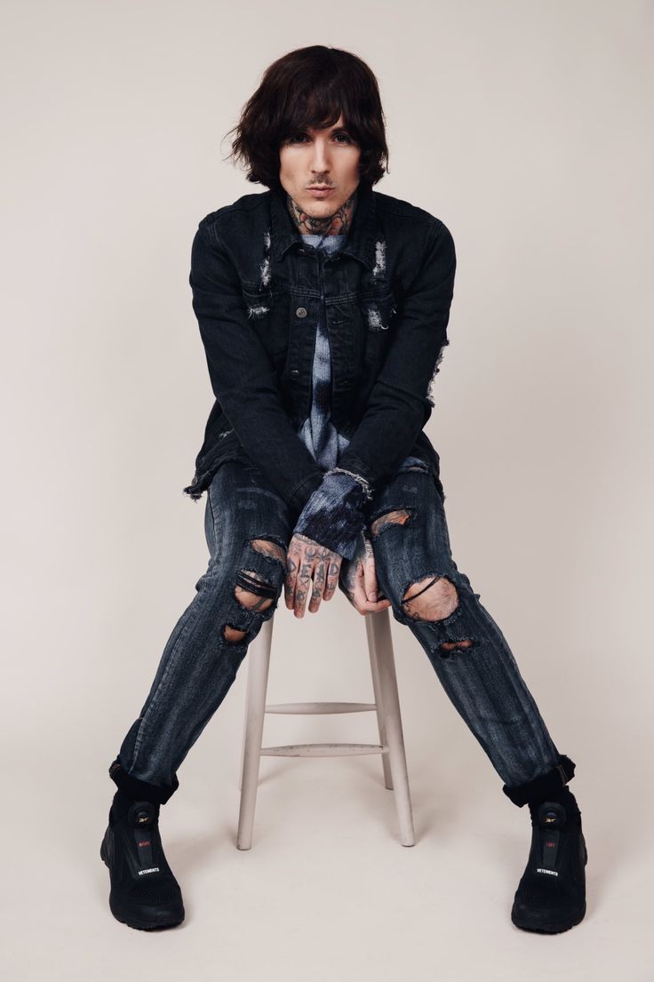 a young man sitting on top of a stool wearing ripped jeans and black boots with his hands in his pockets