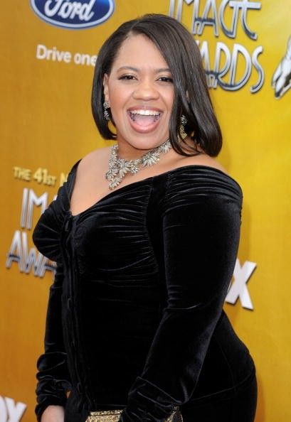 a woman in a black dress smiling on the red carpet at an awards event with her hands in her pockets