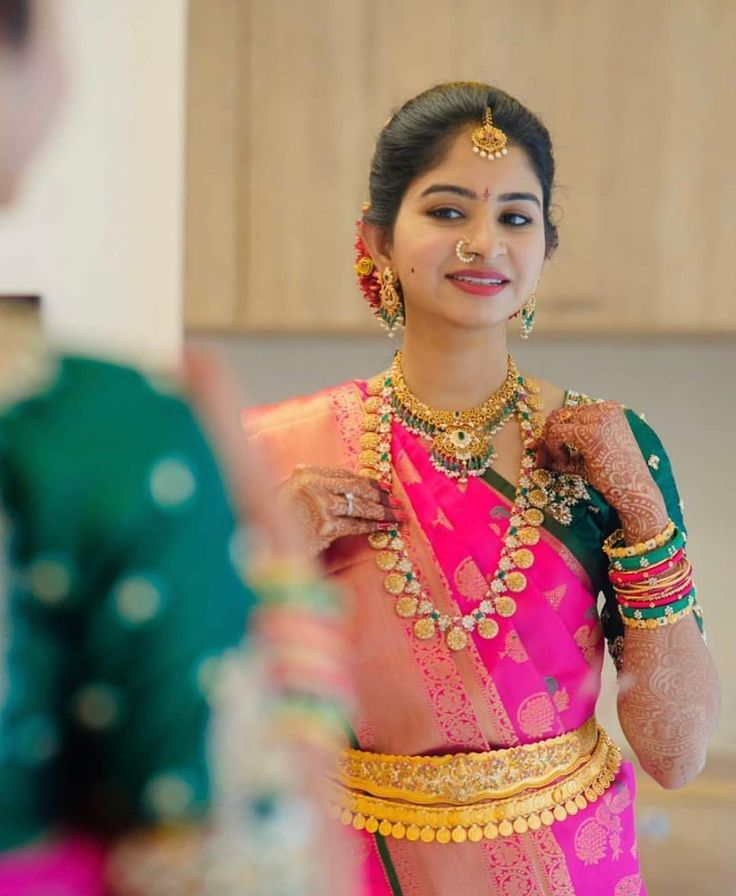 a woman in a pink and green sari with gold jewelry on her neck is looking into the mirror