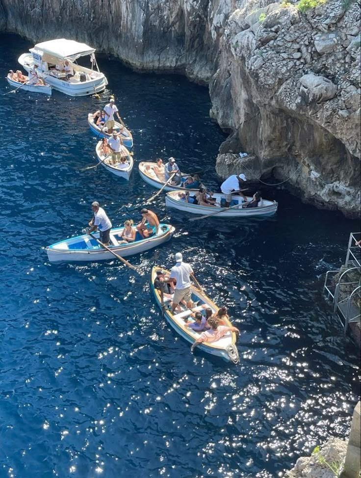 several small boats are in the water with people on them and some cliffs behind them