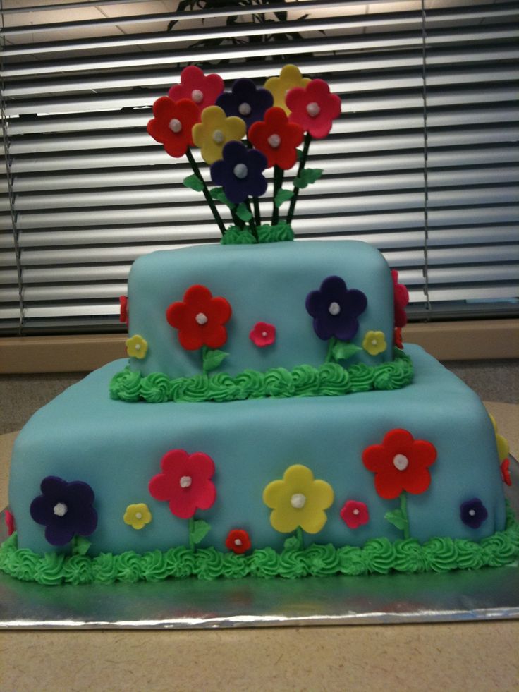 a three tiered blue cake with flowers on the top and green icing, sitting in front of a window