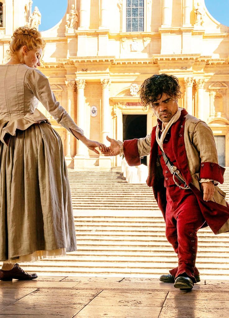 a man and woman dressed in period clothing holding hands while standing on steps with an old building in the background