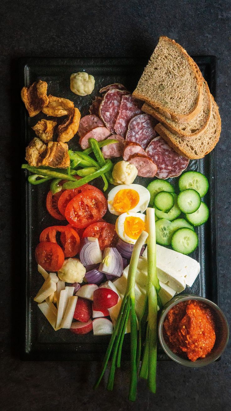 an assortment of meats, vegetables and bread on a black tray with dipping sauce