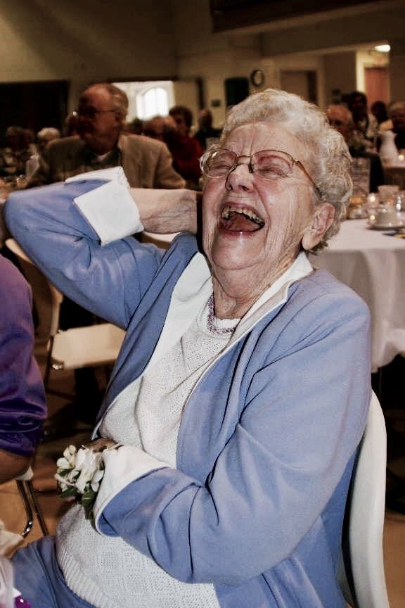 an older woman laughing while sitting in a chair at a table with other people around her