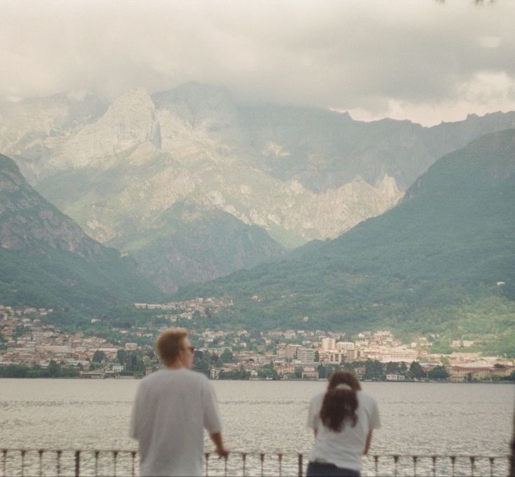 a man and woman standing next to each other near the water with mountains in the background