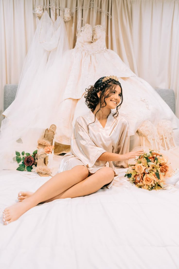 a woman sitting on top of a bed next to a wedding dress and bouquet in her hand