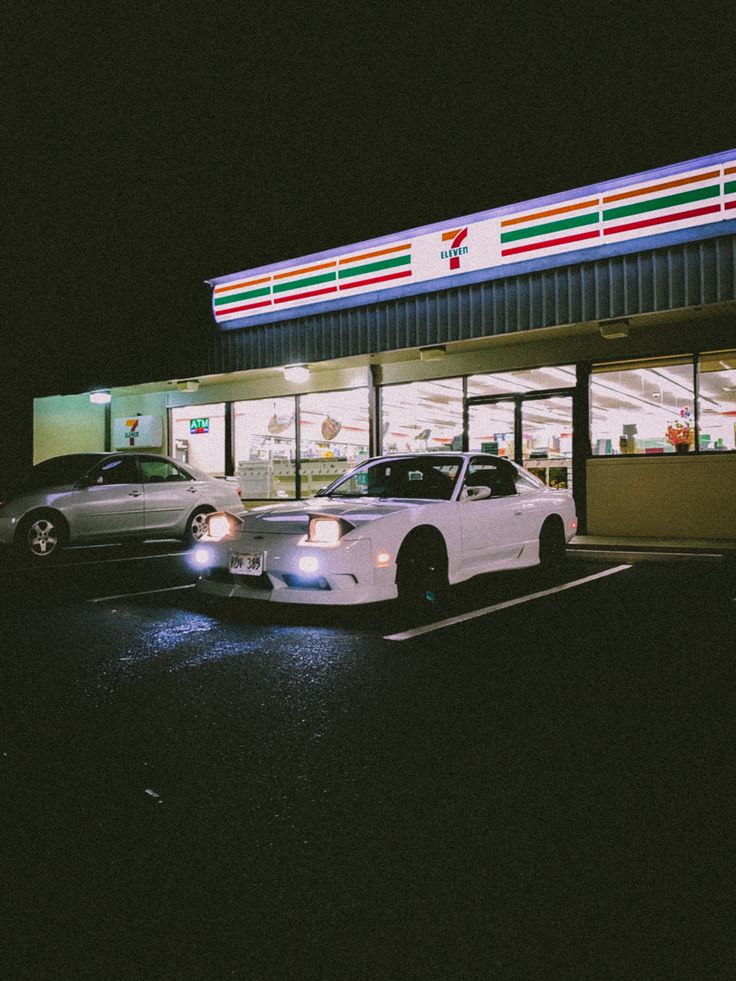 two cars are parked in front of a gas station at night with the lights on