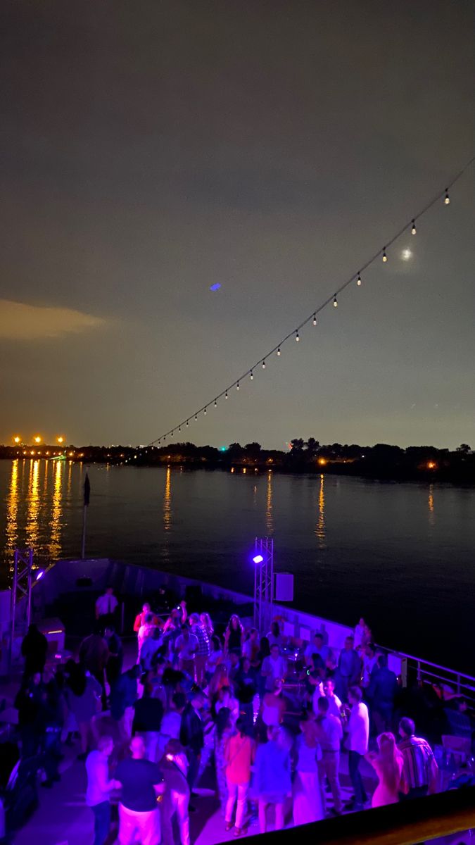 people are gathered on the deck of a boat at night with lights strung across the water