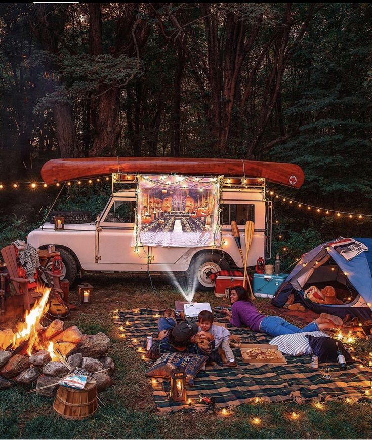 a camper van parked in the woods with its lights on and people sitting around it