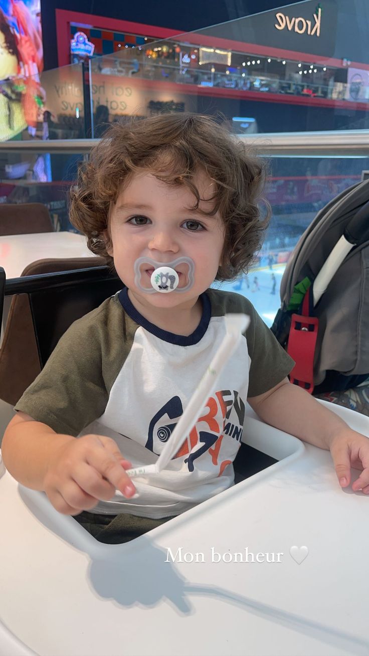 a little boy sitting at a table with a pacifier in his mouth and wearing a t - shirt