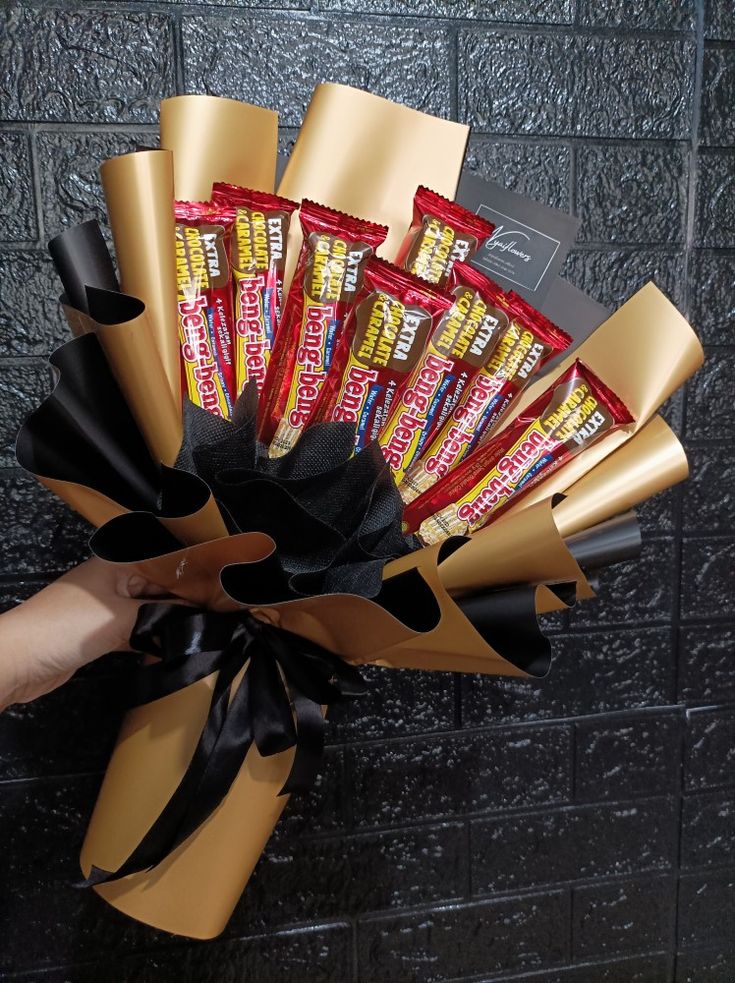 a hand holding a bunch of candy wrapped in gold foil and black ribbon with a brick wall behind it