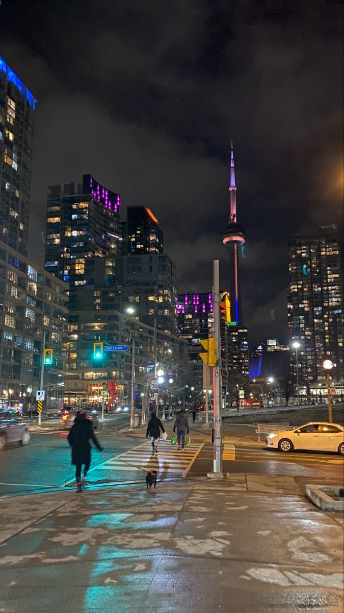 people are walking on the sidewalk in front of some tall buildings at night with bright lights