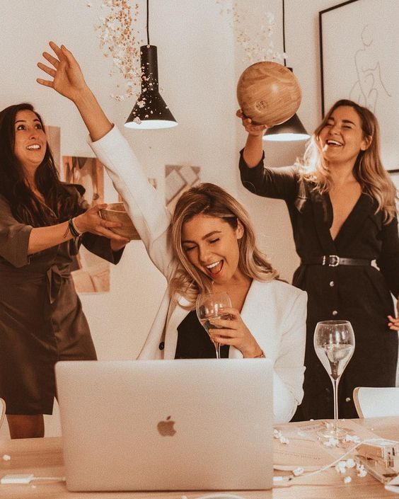 three women at a table with wine glasses in front of a laptop and one holding a wooden ball above her head